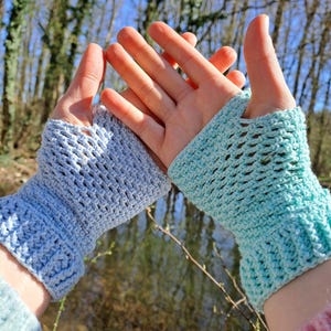 Könnte beinhalten: Zwei gehäkelte Fingerhandschuhe, einer hellblau und einer türkis, werden vor einem verschwommenen Hintergrund aus Bäumen und Wasser gehalten. Die Handschuhe haben ein strukturiertes, offenes Design, perfekt für kühles Wetter.