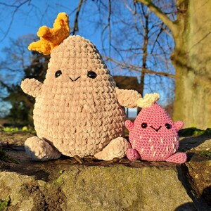 Könnte beinhalten: Zwei handgefertigte Häkeltiere. Eines ist ein großes, beigefarbenes Wesen mit einer gelben Krone. Das andere ist ein kleineres, rosa Wesen mit weißen Akzenten. Beide haben schwarze Augen und lächelnde Gesichter. Sie sitzen auf einem moosbewachsenen Felsen.