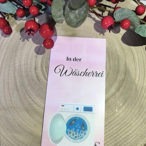 Könnte beinhalten: Eine rosa Karte mit den Worten "In der Wäscherei" und einer Waschmaschinenillustration. Die Waschmaschine hat eine offene Tür mit blauem Wasser und Blasen. Rote Beeren und grüne Blätter sind im Hintergrund.