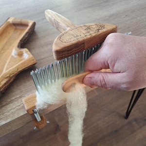 Könnte beinhalten: Ein Holzkamm für Wolle mit dem Text "Woolly Lamb" auf dem Griff. Der Kamm hat Metallzinken und wird zur Verarbeitung von Wolle verwendet. Das Bild zeigt den Kamm in Gebrauch, mit einer Hand, die einen Wollstrang hält.