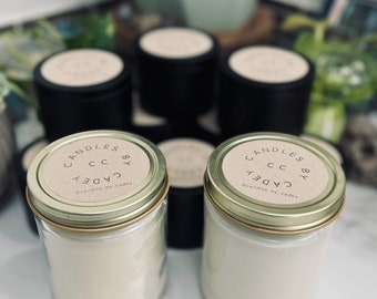 May include: Several candles in glass jars and black tins are arranged on a white surface. The glass jars have gold lids and labels that read "Candles by Cadey." The candles are white and appear to be unscented.