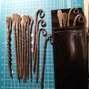 May include: A set of handcrafted metal tools, including twisted and curved designs, displayed on a blue cutting mat. The tools vary in shape, with some having spiral handles and others featuring looped ends. A black leather pouch holds additional tools.