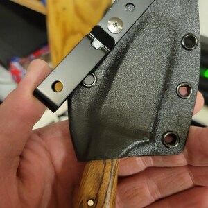May include: A small, black knife sheath with a clip, attached to a wooden handle. The sheath is textured and secured with screws. The handle has a natural wood grain pattern and a small, round, white accent.