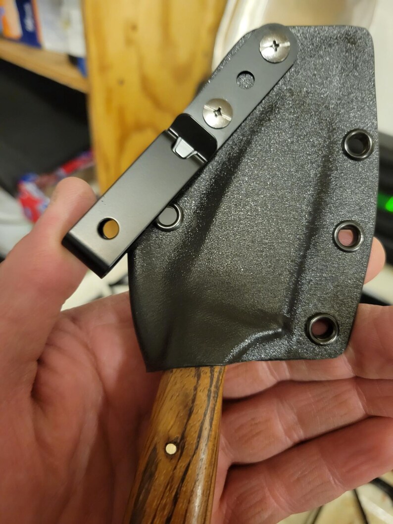 May include: A small, black knife sheath with a clip, attached to a wooden handle. The sheath is textured and secured with screws. The handle has a natural wood grain pattern and a small, round, white accent.