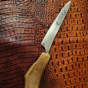 May include: A handmade knife with a brown antler handle and a silver blade. The blade has an intricate design etched into it. The knife is resting on a brown alligator skin background.