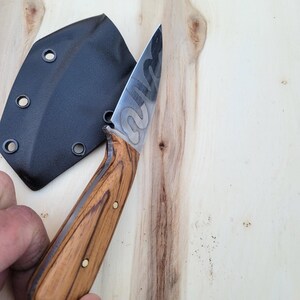 May include: A handcrafted knife with a wooden handle and a patterned blade. The knife is shown with a black sheath. The handle is a light brown color with dark brown graining. The blade has a unique design.