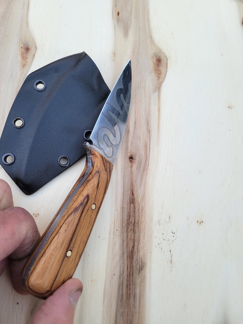 May include: A handcrafted knife with a wooden handle and a patterned blade. The knife is shown with a black sheath. The handle is a light brown color with dark brown graining. The blade has a unique design.