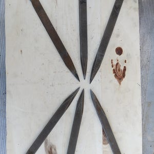 Puede incluir: Siete puntas de metal oscuro y desgastado dispuestas en forma de estrella sobre una superficie de madera clara. Cada punta tiene una cabeza plana y texturizada y un extremo puntiagudo. Algunas puntas tienen una pequeña marca decorativa.