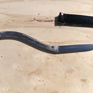 May include: A hand-forged, curved metal knife with a pointed blade and a rounded handle. The knife has a silver finish and is accompanied by a black leather sheath. The knife is displayed on a light-colored wooden surface.