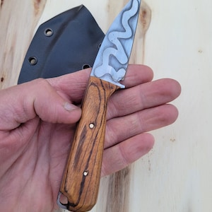 May include: A small, handcrafted knife with a patterned blade and a wooden handle. The blade has a unique, wavy design. The handle is made of wood with a visible grain. A black sheath is visible in the background.