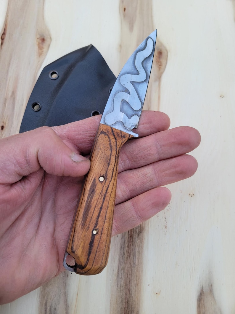 May include: A small, handcrafted knife with a patterned blade and a wooden handle. The blade has a unique, wavy design. The handle is made of wood with a visible grain. A black sheath is visible in the background.