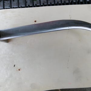 Puede incluir: Una hoja de metal curvada con una base rectangular plana. La hoja es plateada y tiene un extremo afilado y puntiagudo. El objeto es probablemente una herramienta o parte de un equipo más grande. El artículo está sobre una superficie de madera de color claro.