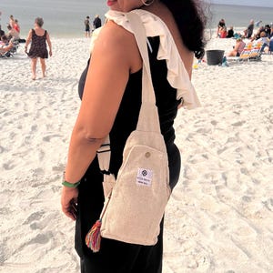 May include: A woman wearing a black dress and a wide-brimmed straw hat with a black band is carrying a beige hemp bag with a tassel. The bag has the text "Style Savvy Hemp Bag" printed on it.