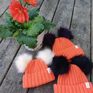 Puede incluir: Cuatro gorros de punto naranja con pompones de piel sintética en blanco, negro y morado. Los gorros son acanalados y tienen una pequeña etiqueta blanca. Una planta en maceta con flores naranjas y hojas verdes está cerca.