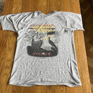 May include: Grey t-shirt featuring a graphic of a bird and a creature with a crown on its head. The text "ROXY MUSIC" and "AVALON" is printed on the shirt.