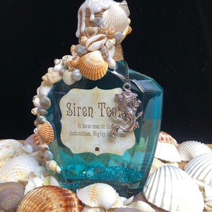 Może przedstawiać: Niebieska szklana butelka z zawieszką w kształcie syreny i etykietą z napisem "Siren Tears. It lures men to their destruction. Highly dangerous." Butelka jest ozdobiona muszelkami i białą, spływającą substancją.