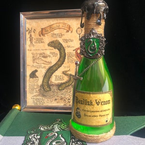 Può includere: Una bottiglia di vetro verde con un sigillo di cera nera sulla parte superiore. La bottiglia è etichettata "Basilisk Venom" e ha un ciondolo d'argento con un serpente verde. La bottiglia è appoggiata su una superficie verde e grigia con una toppa di serpente verde che dice "Slytherin".
