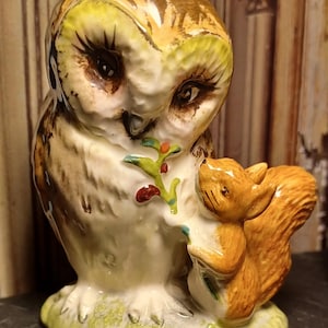 May include: A ceramic owl figurine with brown and white feathers, holding a small orange squirrel figurine with a bushy tail. The owl has large, black eyes and a green branch with red berries in its beak. The base of the figurine is green and brown.