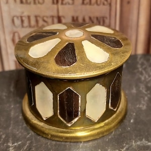 May include: A brass trinket box with a decorative lid. The lid features a flower design with alternating white and brown petals. The box is decorated with a geometric pattern of white and brown.