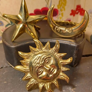 May include: Three gold-toned metal ornaments in the shape of a star, crescent moon, and sun. The sun has a face with eyes, nose, and mouth.