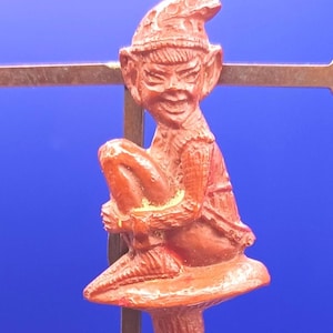 May include: A reddish-brown molded plastic figurine of a pointed-ear creature wearing a cap and sitting on a mushroom. The figure has a textured surface, a wide grin, and is mounted on a thin metal rod against a solid blue background.