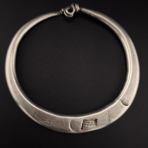 Peut inclure: Un collier rigide argenté de forme circulaire. Le collier est doté d'une fermeture à fermoir et de marques décoratives sur le devant. Le bijou est présenté sur un fond sombre.