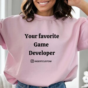May include: A pale pink sweatshirt with the text "Your favourite Game Developer" in black. The sweatshirt also features an Instagram handle below the text. The model is wearing blue jeans.