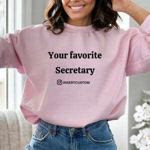 Può includere: Una felpa rosa chiaro con la scritta nera "Your favorite Secretary". La felpa include anche un nome utente Instagram. La modella indossa jeans blu.