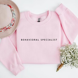 Könnte beinhalten: Ein rosafarbenes Sweatshirt mit Rundhalsausschnitt und dem Aufdruck "BEHAVIORAL SPECIALIST" auf der Vorderseite.