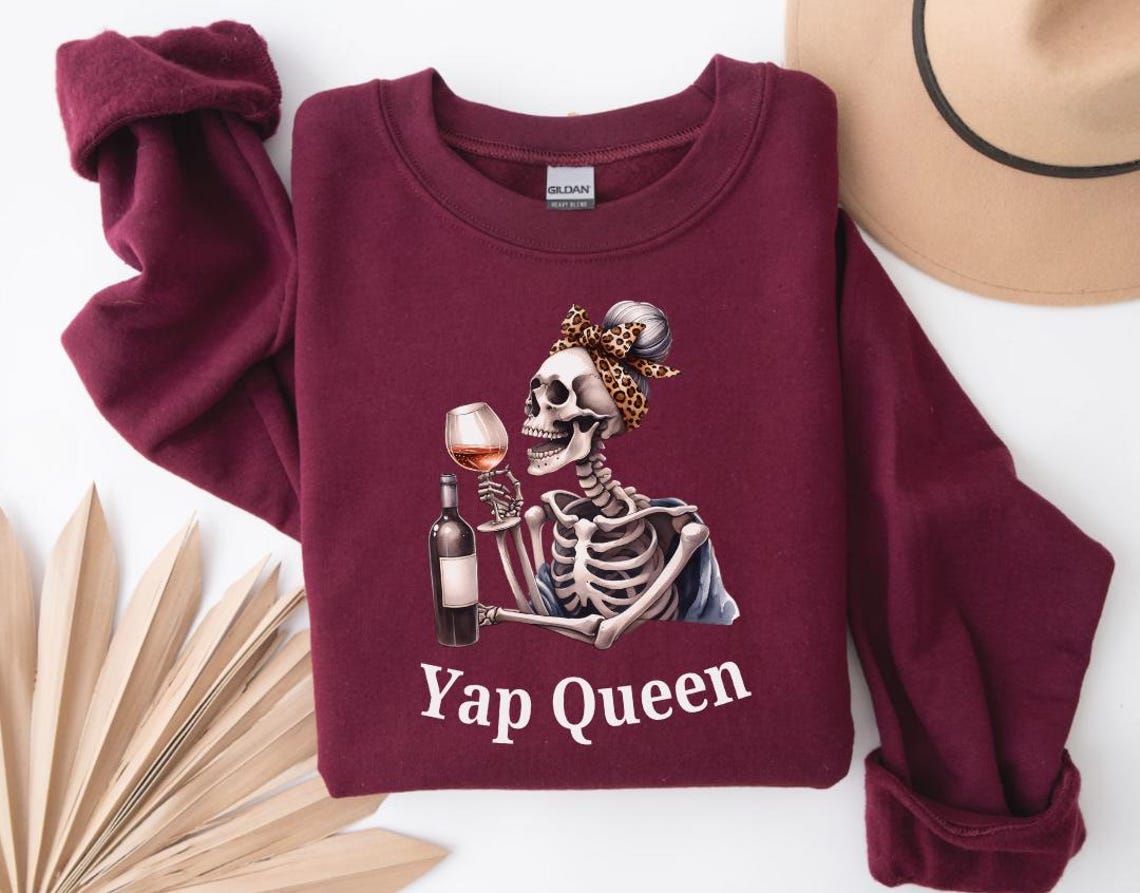 Może przedstawiać: Bordowy sweter z nadrukiem szkieletu trzymającego kieliszek wina i butelkę wina. Szkielet ma na sobie chustkę, a na swetrze widnieje napis "Yap Queen".