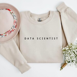 Może przedstawiać: Beżowa bluza z napisem "DATA SCIENTIST" w kolorze czarnym. Na zdjęciu znajduje się również beżowy kapelusz z kwiatową opaską i mały bukiet białych kwiatów. Bluza ma długie rękawy i okrągły dekolt.