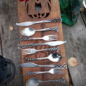May include: A set of handcrafted silverware, including knives, forks, and spoons, arranged on a wooden serving board. The metal utensils have decorative twisted handles. The wooden board has carved details. The overall aesthetic is rustic and artisanal.