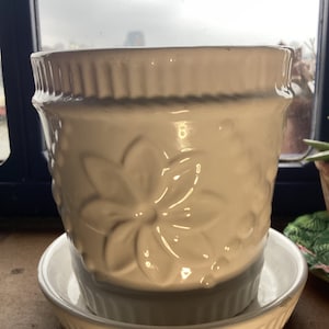 May include: Cream-coloured ceramic plant pot with a matching saucer. The pot has a floral pattern and a ribbed rim. The saucer is round with a ribbed edge. Suitable for indoor or outdoor use.