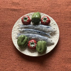 May include: A decorative ceramic plate featuring sardines, tomatoes, and bell peppers. The sardines are painted in shades of blue and grey, while the tomatoes are red and the bell peppers are green. The plate has a textured, white surface.