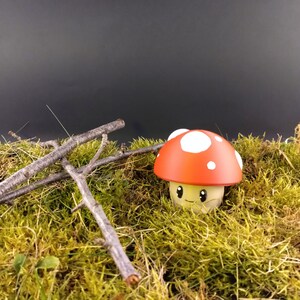 Peut inclure: Une figurine en forme de champignon avec un chapeau rouge et des taches blanches, posée sur de la mousse verte et des brindilles. La figurine a un visage souriant et un pied de couleur crème. Thème : nature et fantaisie.