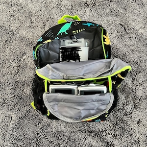 May include: A black backpack with a dinosaur print and green accents. The backpack is open and has two small electronic devices inside.