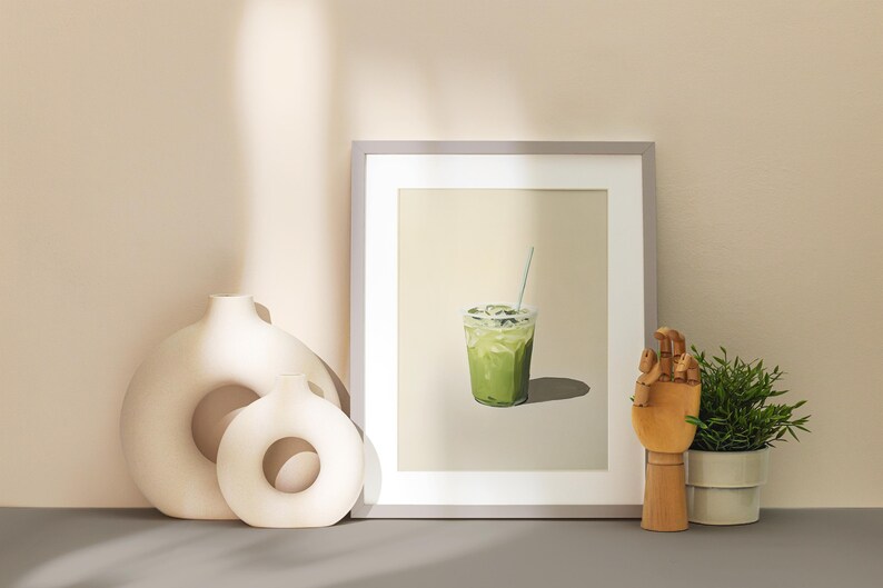 May include: A framed print of a green iced drink with a straw in a clear plastic cup. The print is on a beige background and is framed in a light gray frame.