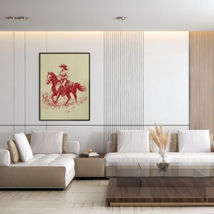 May include: Framed art print featuring a red illustration of a person riding a horse. The artwork is set against a cream-colored background and framed in black. The image is displayed in a modern living room setting.