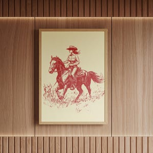 May include: Framed print featuring a red and cream illustration of a cowgirl riding a horse. The cowgirl wears a hat and is seated on a horse galloping through a field of grass. The artwork is in a wooden frame and mounted on a wood-paneled wall.