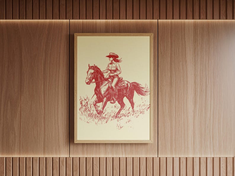 May include: Framed print featuring a red and cream illustration of a cowgirl riding a horse. The cowgirl wears a hat and is seated on a horse galloping through a field of grass. The artwork is in a wooden frame and mounted on a wood-paneled wall.