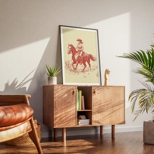 May include: A framed print featuring a red and white illustration of a person riding a horse. The artwork is displayed on a wooden cabinet with books and a small plant. A leather chair and a potted plant are also in the room.