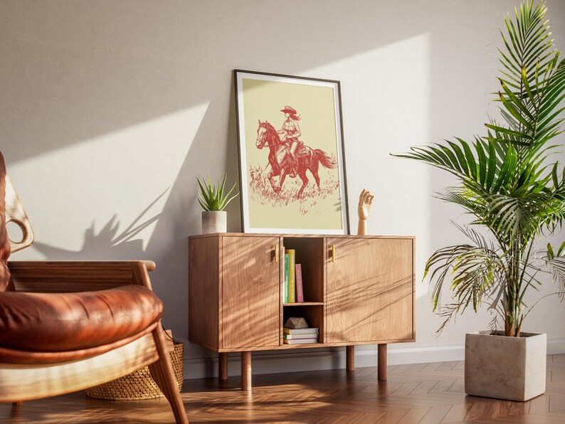 May include: A framed print featuring a red and white illustration of a person riding a horse. The artwork is displayed on a wooden cabinet with books and a small plant. A leather chair and a potted plant are also in the room.