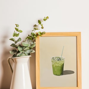 May include: A framed print of a green iced drink with a straw in a clear glass. The print is on a light beige background and is framed in a natural wood frame.