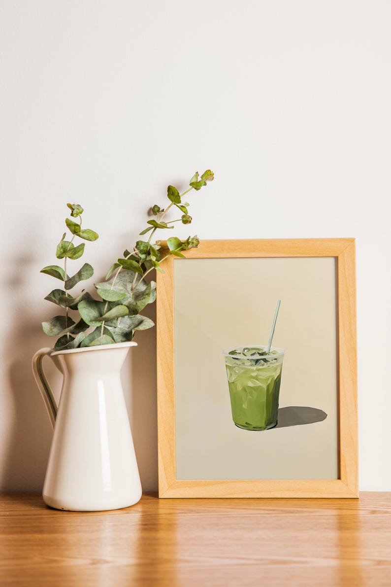 May include: A framed print of a green iced drink with a straw in a clear glass. The print is on a light beige background and is framed in a natural wood frame.