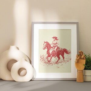 May include: Framed print of a cowgirl riding a horse, rendered in red tones against a cream background. The artwork is displayed with decorative vases, a wooden hand sculpture, and a potted plant. The overall theme is Western.
