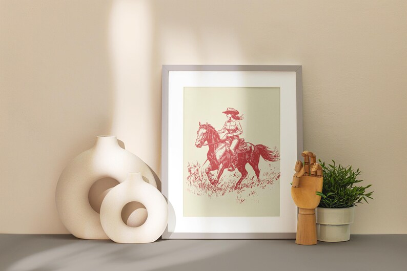 May include: Framed print of a cowgirl riding a horse, rendered in red tones against a cream background. The artwork is displayed with decorative vases, a wooden hand sculpture, and a potted plant. The overall theme is Western.