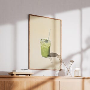 May include: A framed print of a green iced drink with a straw in a clear plastic cup. The background is a light yellow color.
