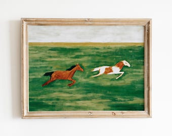 May include: A framed painting depicts two horses galloping across a green field. One horse is brown, the other is white and brown. The sky is a pale gray, and the frame is a light wood color.