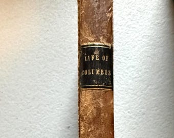 May include: An antique book titled "Life of Columbus." The book has a worn, brown leather cover with a black label on the spine. The text on the label is in gold lettering. The book's spine shows signs of age and wear.