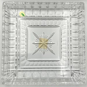 May include: Square, clear glass dish with a starburst design in the center. The dish has a multi-layered design with a textured border and a small, round sticker in the upper left corner.